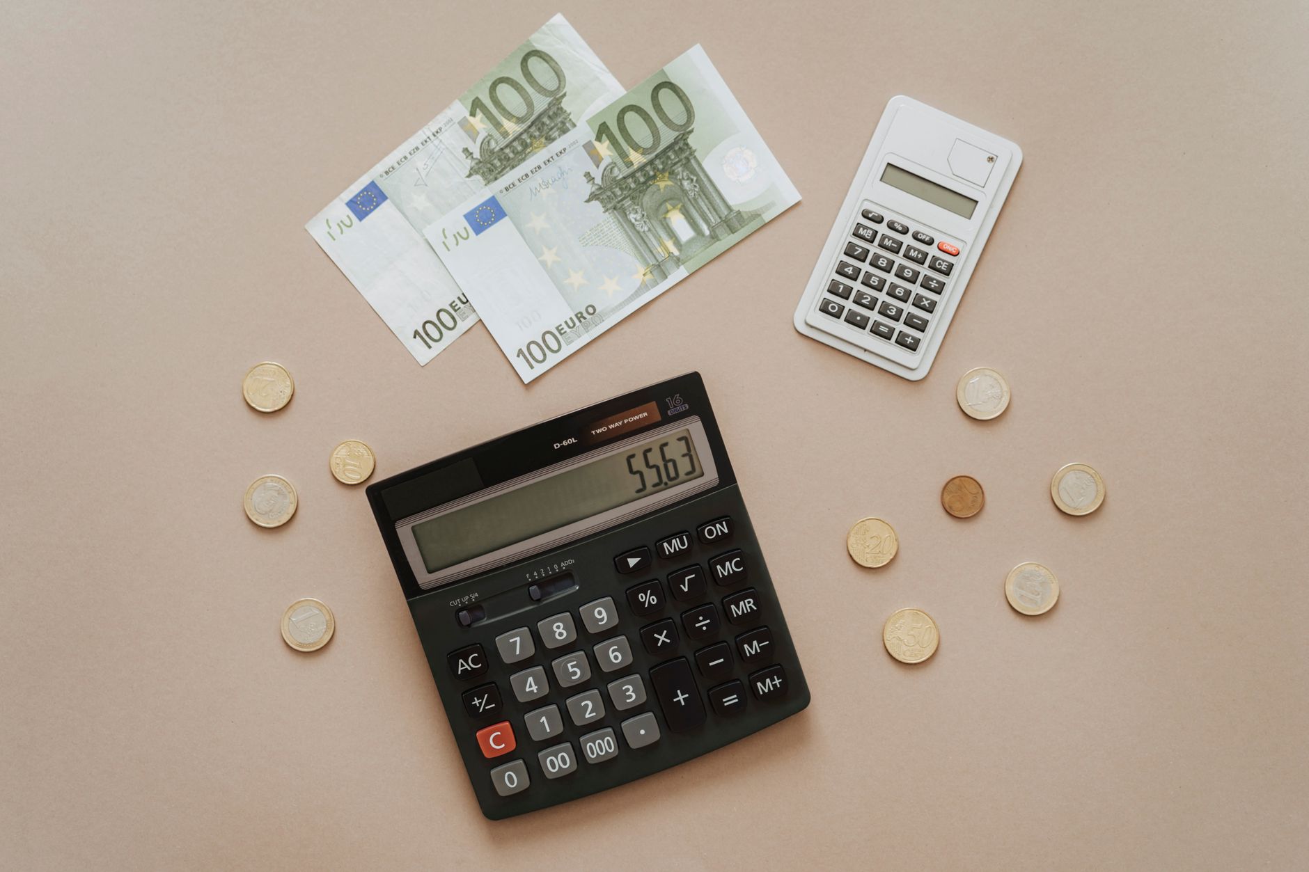 Calculatrice noire, petite calculatrice blanche, deux billets de 100 euros et pièces de monnaie posés sur un fond beige, vus de dessus.
