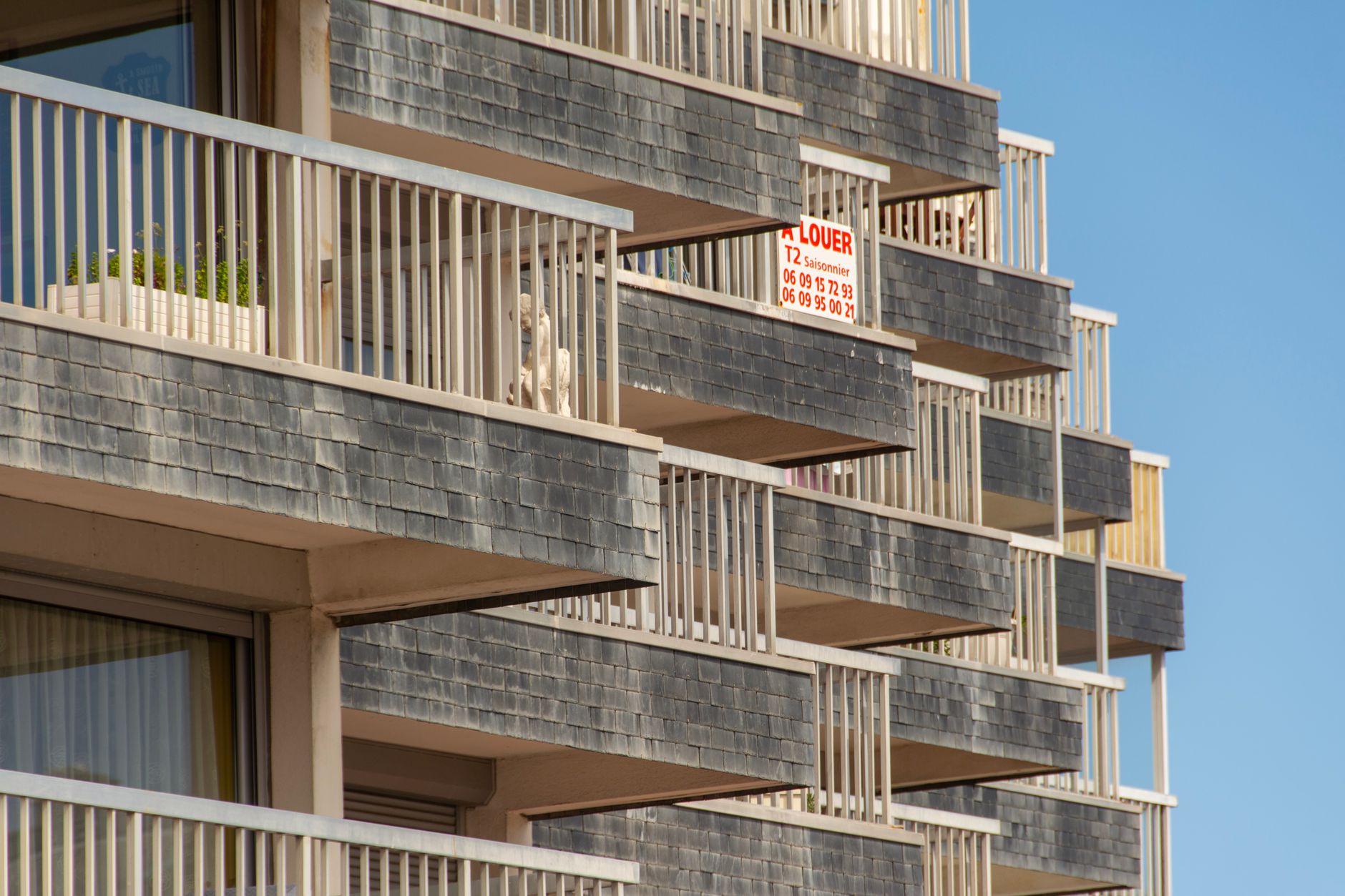 Façade d’immeuble à balcons en ardoise grise, avec un petit panneau « À LOUER » accroché à un garde-corps, sous un ciel bleu.