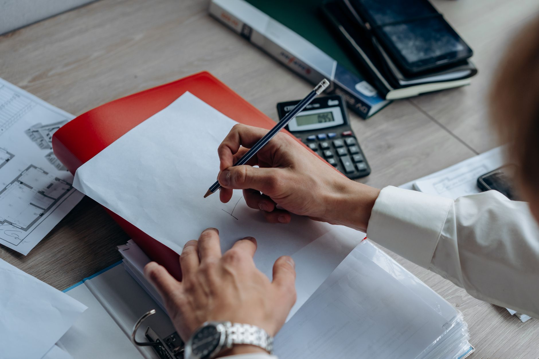 Mains d’une personne écrivant avec un crayon sur une feuille posée sur un classeur, près d’une calculatrice sur un bureau.